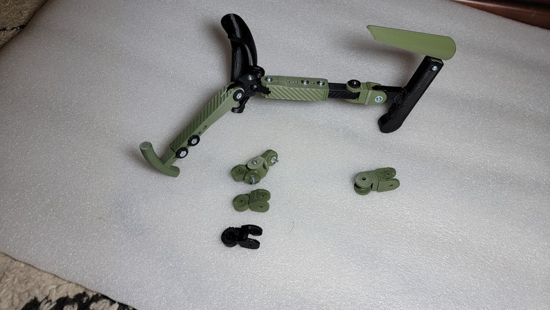 May include: A green and black 3D printed articulated robotic arm with multiple joints. The arm is disassembled and laid out on a white surface. The arm is made of plastic and has a carbon fibre pattern.