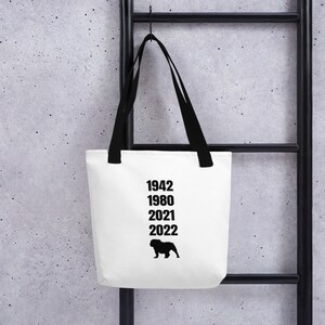 Op de afbeelding: Witte tote bag met zwarte handvatten en zwarte tekst die "1942 1980 2021 2022" luidt en een zwart silhouet van een bulldog.