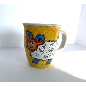 May include: A ceramic mug with a yellow background and a cartoon sheep design. The sheep has a red face, blue horns, and white wool with black swirls. The mug has a white handle and a gray rim.
