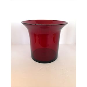 May include: A ruby red glass vase with a flared top and a cylindrical body. The vase has a smooth, glossy finish and a dark base. The interior of the vase is visible, and the glass appears to be thick and sturdy. The vase is likely used for decorative purposes.