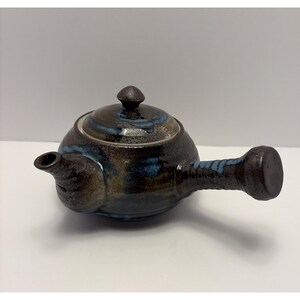 May include: A ceramic teapot with a dark brown body, a lid, and a long, cylindrical handle. The teapot has blue and gold accents. The spout is on the left side. The lid has a small knob on top.