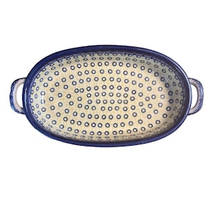 May include: Oval ceramic baking dish with a blue rim and handles. The interior has a cream background with a pattern of small blue circles. The dish is designed for baking and serving.
