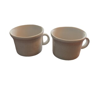 May include: Two off-white ceramic mugs with rounded bodies and small handles. The mugs have a slightly textured surface and a simple, classic design. The interior of the mugs is a lighter shade of white.