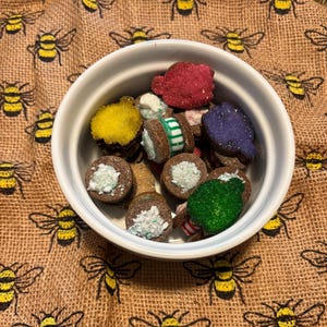 May include: A white ceramic bowl filled with colorful, decorated cookies. The cookies are various shapes and colors, including red, yellow, green, and purple. The bowl sits on a burlap cloth with a repeating pattern of yellow and black bees.