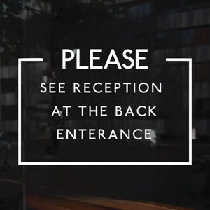 May include: A rectangular sign with a white border on a dark background featuring the white text PLEASE SEE RECEPTION AT THE BACK ENTERANCE. The sign is designed for clear communication and is displayed on a glass surface with a dark, blurred interior.