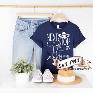 May include: A navy blue t-shirt with white text that reads "Next Stop Isla Mujeres" with a sombrero, margarita glass, and airplane graphic. The shirt is on a hanger with a pair of blue jeans, white sneakers, and a straw hat.