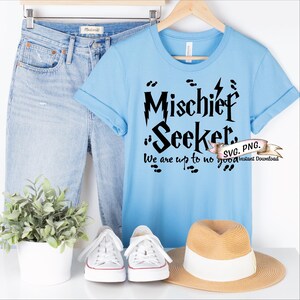 Może przedstawiać: Jasnoniebieska koszulka z czarnym napisem "Mischief Seeker. We are up to no good." Tekst jest w stylizowanym foncie i zawiera grafikę błyskawicy. Koszulka znajduje się na białym tle z dżinsami, kapeluszem i butami sportowymi.
