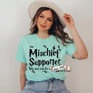 May include: A woman wearing a light blue t-shirt with the text "Mischief Supporter" in black. The text is in a stylized font with lightning bolts and footprints. The text "We are up to no good" is also included on the shirt.