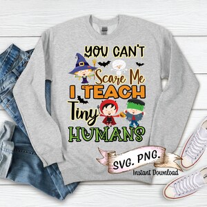 May include: Gray long-sleeve sweatshirt with the text "You Can't Scare Me I Teach Tiny Humans" and a graphic of four children dressed in Halloween costumes. The text "SVG. PNG. Instant Download" is also included.