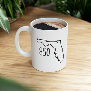 May include: White ceramic coffee mug with a black outline of the state of Florida and the number 850 printed on it. The mug is filled with coffee.