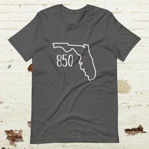Puede incluir: Una camiseta gris oscuro con un contorno blanco del estado de Florida y el número 850 impreso en la parte delantera.