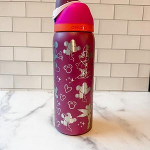 A pink and purple children's sippy cup with a Disney-themed design, including Mickey Mouse and Minnie Mouse characters, is sitting on a marble countertop.