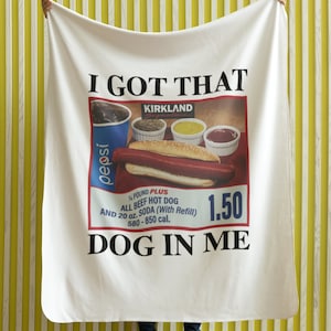 May include: White blanket with the text "I GOT THAT" and a picture of a hot dog, a soda, and condiments. The text "KIRKLAND" is above the picture. The text "¼ POUND PLUS ALL BEEF HOT DOG AND 20 oz. SODA (With Refill) 580-850 cal." is below the picture. The text "1.50" is to the right of the picture. The text "DOG IN ME" is below the picture.