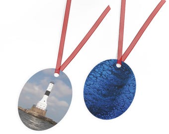 May include: Two oval-shaped hanging ornaments with red ribbon loops. One ornament features a white and black lighthouse on a rocky base surrounded by water under a cloudy sky. The other ornament displays a dark blue abstract fluid paint pattern.