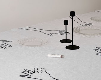 May include: A white tablecloth with black embroidered hand designs. Two black metal candle holders are on the table, one taller than the other. A white lighter is on the table.
