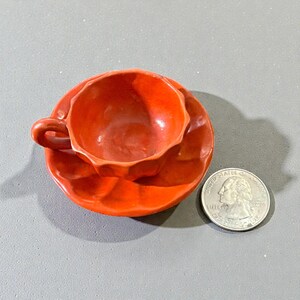 May include: A small, orange ceramic teacup and saucer set. The cup has a handle and a faceted design, sitting on a matching saucer. A U.S. quarter is next to the set for size comparison. The set is on a gray surface.