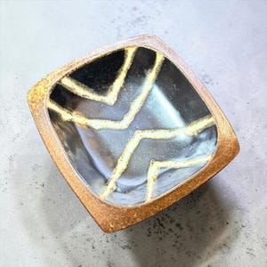 May include: A square ceramic bowl with a textured, metallic copper-colored rim and a glossy black interior. The interior features two distinct, parallel yellow lines that form a chevron pattern, creating a sharp contrast against the dark surface.