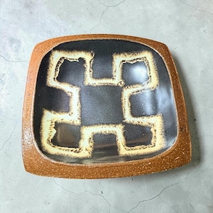 May include: A square ceramic dish with rounded corners, featuring a textured, speckled brown rim and a smooth, dark charcoal-colored center. The interior is decorated with a bold, cream-colored geometric pattern that resembles a stylized cross.