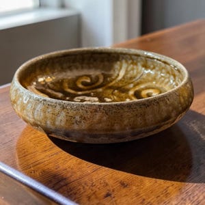May include: A small, handcrafted ceramic bowl with a rounded shape. The bowl features a textured, earthy brown exterior and a glossy, swirling pattern in the interior. The bowl is sitting on a wooden surface.