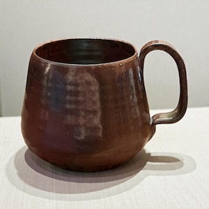 May include: A handmade, brown ceramic mug with a rounded body and a sturdy handle. The mug has a rustic, textured appearance with variations in the glaze, suggesting a unique, artisanal quality. The mug is designed for hot beverages.