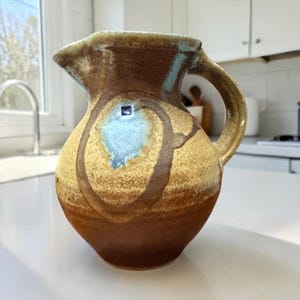 May include: A handmade ceramic pitcher with a rounded body and a handle. The pitcher features a color palette of brown, tan, and blue, with a unique design. The pitcher is suitable for serving beverages and adds a rustic touch to any kitchen.