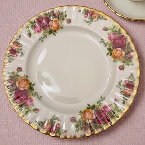 May include: A round porcelain plate with a scalloped edge and a gold-trimmed rim. The plate features a cream-colored center and a border decorated with clusters of red and yellow roses with green leaves arranged around the perimeter.