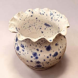 May include: A ceramic bowl with a rounded body and a wide, ruffled, scalloped rim. The surface is finished in a glossy white glaze featuring a speckled pattern of dark blue paint splatters distributed across the interior and exterior.