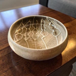 May include: A round ceramic bowl with a speckled beige and olive green glaze. The interior features four vertical, wavy white lines, each decorated with a series of small, dark brown dots arranged in a linear pattern along the center of the shapes.