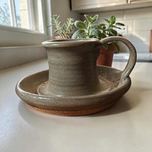 May include: A handmade ceramic candle holder with a wide, flat circular base and a central cylindrical cup. The piece features a muted, earthy taupe glaze with a smooth finish, while the bottom edge reveals the raw, unglazed terracotta clay.
