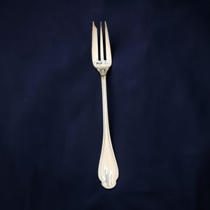 May include: A silver fork with three tines, a long handle, and decorative detailing. The fork is set against a dark blue background, highlighting its polished surface and elegant design. This utensil is suitable for formal dining.