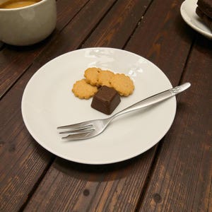 May include: A white plate holds a small assortment of treats: three golden-brown, flower-shaped cookies and a dark chocolate square. A silver fork rests beside the sweets. A cup of coffee is in the background. The scene is set on a dark wooden table.