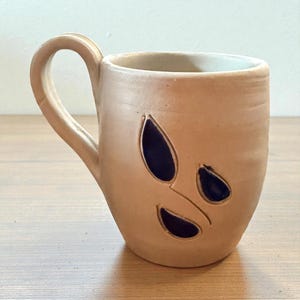 May include: A light tan ceramic mug with a single handle and three dark blue, teardrop-shaped cutouts on the front. The mug has a smooth, matte finish and a cylindrical body with a slightly tapered base, showcasing a simple, functional design.