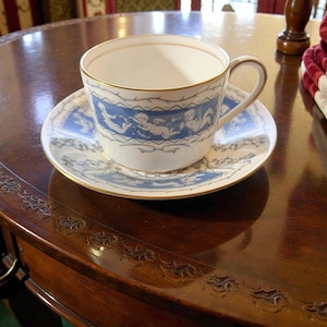 May include: White teacup and saucer set with a gold rim and a blue and white cherub design. The teacup has a curved handle and sits on a matching saucer. The set is placed on a dark wooden table.