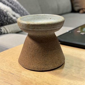 May include: A handcrafted ceramic vessel featuring a wide, shallow bowl and a conical base. The bowl is off-white, and the base has a textured brown finish. The item is placed on a wooden surface, possibly for display or use.