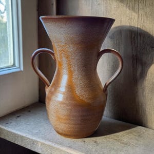 May include: A ceramic vase with two curved handles and a wide, flared rim. The surface features a mottled glaze with blended tones of warm amber, burnt orange, and creamy white, creating a textured appearance across the rounded body.