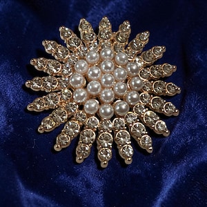 May include: A circular brooch featuring a central cluster of white faux pearls surrounded by two radiating layers of clear, faceted rhinestones set in a gold-toned metal frame, creating a sunburst pattern with a polished, reflective finish.