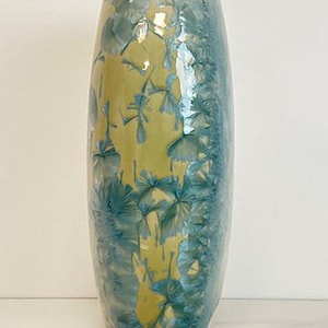 May include: A tall, ceramic vase with a unique crystalline glaze. The vase features a vertical design with a yellow center and blue-green edges. The glaze has a textured, almost floral pattern. The vase is approximately 12 inches tall.