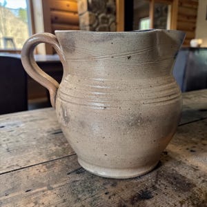 May include: A handmade, beige ceramic pitcher with a curved handle. The pitcher has a wide base, a rounded body, and a slightly flared rim. Horizontal lines and subtle markings add texture to the surface. The pitcher sits on a wooden surface.