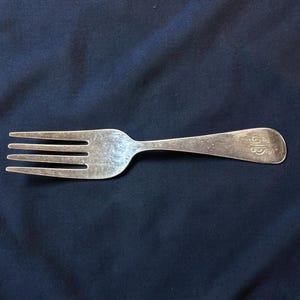 May include: A silver fork with four tines, resting on a dark blue surface. The handle has a rounded end with an engraved design. The fork is a classic piece of silverware.