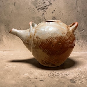 May include: A handmade ceramic pitcher with a unique design. The pitcher features a spout, two small handles, and a rounded body. The color is a blend of off-white and reddish-brown, with visible texture. The pitcher is approximately 6 inches tall.