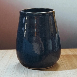 May include: A dark blue ceramic vase with a glossy finish. The vase has a rounded body that tapers slightly towards the top. The interior is dark, and the base is unglazed. The vase sits on a wooden surface.