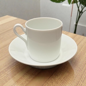 May include: A white ceramic teacup and saucer set. The cup has a curved handle and sits on a matching saucer. The set is placed on a light-colored wooden surface. The cup and saucer are both white.