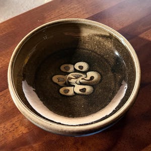 May include: A handmade ceramic bowl with a dark brown interior and a lighter brown exterior. The bowl features a unique design of cream-colored, abstract shapes in the center. The rim is a lighter shade of brown. The bowl is likely used for serving food or as a decorative piece.