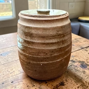 May include: A cylindrical ceramic canister with a textured, ribbed exterior in shades of earthy brown and tan. The vessel features a matching circular lid with a small central knob, showcasing a rustic, hand-thrown appearance with visible glaze variations.