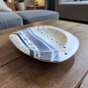 May include: An oval ceramic dish with a cream-colored base featuring a decorative border of blue and gray geometric patterns, including star-like motifs and cracked textures, alongside small green and brown painted accents on the interior surface.