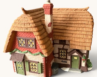Può includere: Una casa in miniatura in ceramica con un tetto di paglia beige testurizzato, pareti in mattoni rossi e sezioni in stucco color crema. La struttura presenta porte verdi, persiane verdi, finestre a traliccio e la parola Cobb sulla parete.