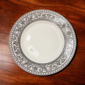 May include: A round ceramic plate with a cream-colored center and a wide border featuring a detailed gray pattern of mythical creatures, foliage, and urns, finished with a thin dotted line along the outer and inner edges of the rim.