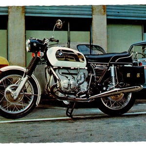 May include: A vintage BMW motorcycle featuring a white fuel tank with black accents, a black leather seat, and chrome exhaust pipes. The motorcycle is equipped with wire-spoke wheels, a round headlight, a single side mirror, and black saddlebags.
