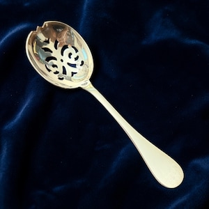 May include: A silver serving spoon with a slotted, oval bowl and a long, slender handle. The bowl features an intricate, openwork design. The spoon is set against a dark blue velvet background, highlighting its polished surface.