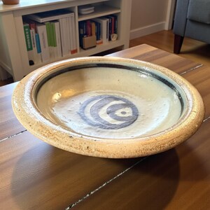 May include: A shallow, round ceramic bowl with a wide, textured, beige-colored rim. The interior of the bowl features a smooth, off-white glaze with a dark blue, hand-painted spiral design centered at the bottom and a dark blue band along the inner edge.